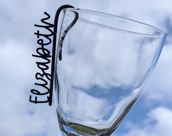 May include: Black acrylic wine glass charm with the name "Elizabeth" written on it. The charm is attached to a wine glass.