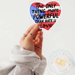 May include: A heart-shaped sticker featuring the Puerto Rican flag design with the text "The ONLY THING more POWERFUL THAN HATE is Love." The sticker is held up by a hand with rings. The sticker is white, red, and blue.