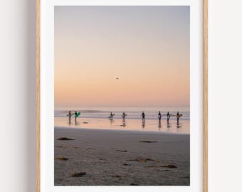 May include: A framed photograph of a beach scene at sunset. Silhouetted figures with surfboards walk along the shoreline toward the ocean. The sky transitions from orange to blue, reflecting on the wet sand.