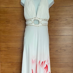 Puede incluir: Vestido maxi blanco con escote pronunciado y cinturón de cuentas doradas. El vestido presenta un diseño floral rojo y dorado en la falda.