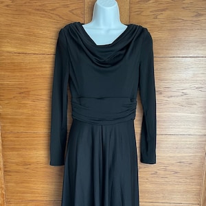 May include: A long, black dress with a cowl neckline and long sleeves. The dress has a gathered waist and a flowing, floor-length skirt. The dress is displayed on a white mannequin against a wooden background.