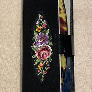 May include: Black case with a floral needlepoint design. The needlepoint features pink, purple, yellow, and green flowers. The case is open, revealing a tortoiseshell-patterned object.