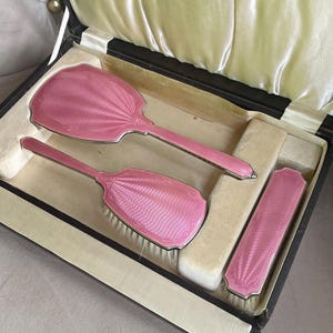 May include: A vintage vanity set featuring a hand mirror, a hairbrush, and a clothes brush, all finished in a vibrant pink enamel with a scalloped shell pattern and silver-toned edges, presented in a rectangular case with a cream satin lining.