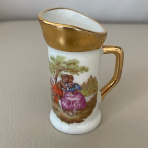 May include: A small white porcelain pitcher with a gold rim and handle. The pitcher features a painted scene of a couple in a garden setting.