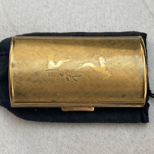 May include: A rectangular, gold-toned compact case with a decorative design of two deer. The compact is set on a black fabric pouch. The case has a clasp closure and a textured surface.