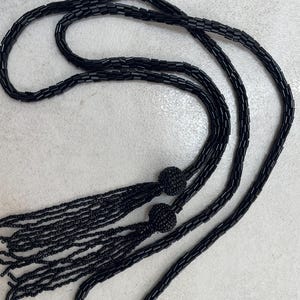 Puede incluir: Un collar largo de color negro compuesto por cuentas cilíndricas ensartadas, con dos detalles de cuentas esféricas que dan paso a múltiples flecos de cuentas colgantes en los extremos, formando un diseño de bucle continuo.