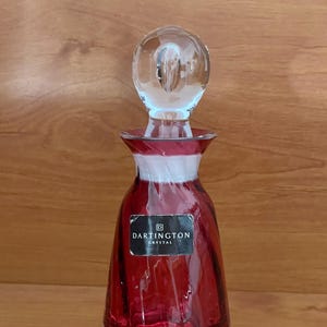 May include: A red glass bottle with a clear glass stopper. The bottle has a label that reads "Dartington Crystal".