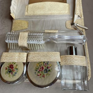May include: Vintage cream-colored travel vanity case with a zipper closure. The case contains a mirror, a hairbrush, two compacts with floral embroidery, and a perfume bottle. The case is made of textured material.