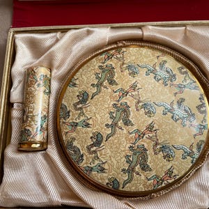 May include: A vintage vanity set comprising a compact mirror and lipstick tube, both decorated with a repeating pattern of mythical creatures in green, blue, and red on a cream backdrop. The set is presented in a gold-edged box with a satin lining.