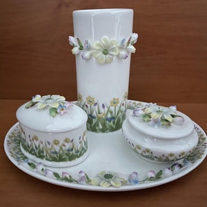 Könnte beinhalten: Ein weißes Porzellan-Schminkset mit Blumenmuster. Das Set enthält ein Tablett, eine Vase und zwei kleine Dosen mit Deckel. Die Blumen sind in Rosa-, Gelb- und Blautönen bemalt.