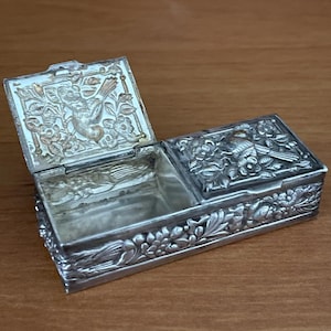 Puede incluir: Una caja rectangular de metal plateado con dos tapas con bisagras. El exterior y el interior de las tapas presentan patrones florales y motivos de aves en relieve. Una tapa está abierta, revelando un compartimento hueco.