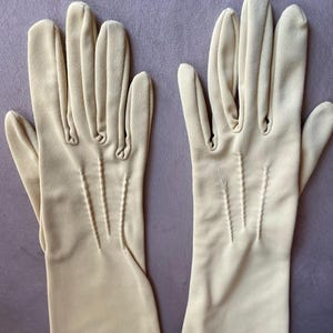 May include: A pair of cream-coloured gloves. The gloves are made of a smooth fabric and feature decorative stitching along the fingers and the back of the hand. The gloves are long, extending past the wrist.