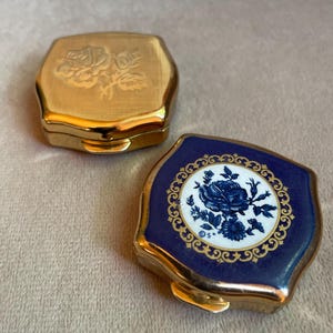 Puede incluir: Dos pastilleros metálicos hexagonales pequeños. Uno es de tono dorado con un diseño de rosa grabado en la tapa. El otro presenta un borde de esmalte azul profundo, una incrustación circular blanca central con un patrón de rosas azules.