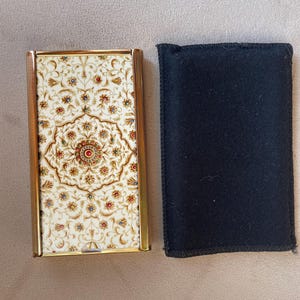 Op de afbeelding: Een rechthoekig sigarettendoosje van goudkleurig metaal met een ingewikkeld crèmekleurig bloemmotief met kleine rode, blauwe en groene accenten, vergezeld van een eenvoudige rechthoekige beschermhoes van zwart vilt met gestikte randen.