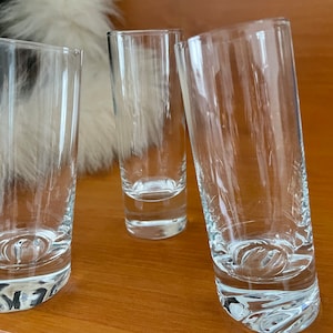 May include: Three clear glass shot glasses on a wooden surface. The glasses are empty and have a rounded bottom.