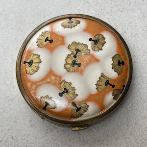 May include: A round, vintage-style compact case with a metallic brass-toned rim. The lid features a decorative pattern of white, cloud-like circular shapes set against a vibrant orange background, accented with small, gold-toned floral motifs.