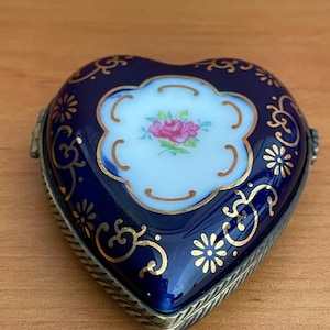 May include: A heart-shaped trinket box with a deep blue enamel finish. The lid features a white floral design with a pink rose at the center, accented by gold filigree and floral patterns. The box has a silver-toned metal frame.