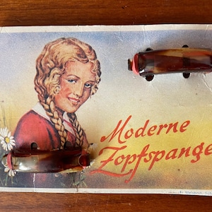 Puede incluir: Tarjeta vintage que anuncia pinzas para el pelo "Moderne Zopfspange". La tarjeta muestra a una joven con trenzas y dos pinzas para el pelo marrones.