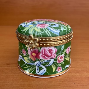 Puede incluir: Una pequeña caja de baratijas redonda verde con un diseño floral. La caja presenta rosas rosas, lazos azules y adornos dorados. La tapa y los lados están decorados con un patrón repetido de flores y lazos.