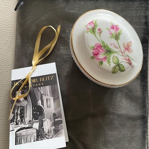 Puede incluir: Un plato de porcelana blanca con borde dorado y un diseño floral con rosas rosas y hojas verdes. El plato está sobre una bolsa de malla negra con una cinta dorada.