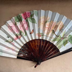 May include: A folding hand fan featuring a light blue background with a floral pattern of pink and peach roses, green leaves, and a small orange butterfly. The fan has a dark brown, tortoiseshell-patterned frame with a glossy finish and a metal rivet.