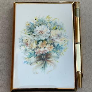 May include: A vintage gold-toned compact with a floral bouquet design on a white background. The compact has a matching gold pen attached to the side. The bouquet features white, peach, and blue flowers with a ribbon.