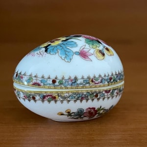 Könnte beinhalten: Eiförmige Schmuckdose in Weiß mit Klappdeckel. Verziert mit farbenfrohen Blumen- und Blättermotiven in Blau, Gelb, Rosa und Grün. Ein goldfarbener Rand akzentuiert Deckel und Boden.
