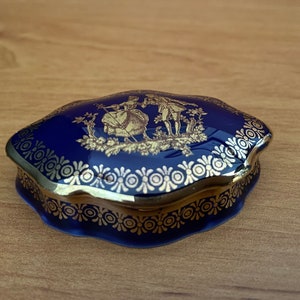 May include: A blue and gold porcelain trinket box with a decorative design of a couple in a garden setting.