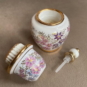 May include: A white ceramic perfume bottle with a gold rim and floral design. The bottle is decorated with pink and purple flowers and green leaves. The bottle's components include a lid with a brush and a stopper.