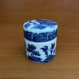 May include: A small, round, white ceramic trinket box with a lid. The box and lid feature a blue and white Willow pattern design depicting a landscape scene. The box is cylindrical with a matching lid.