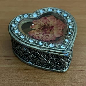 May include: Heart-shaped trinket box with a clear top, showcasing a dried flower. The box is framed with small, clear crystals and has an ornate, bronze-colored metal base. The box is likely used for storing small jewelry items.