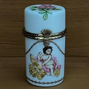 May include: A white porcelain trinket box with a gold hinged lid. The box features a painted image of a cherub sitting on a bundle of wheat, surrounded by a barbed wire design. The lid is decorated with a floral design of pink roses and green leaves.