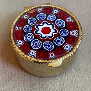 Può includere: Un portapillole rotondo in metallo color oro con un inserto in vetro millefiori sul coperchio. Il design in vetro presenta motivi floreali rossi, bianchi e blu disposti in un motivo circolare. La base in metallo ha un motivo a volute inciso.