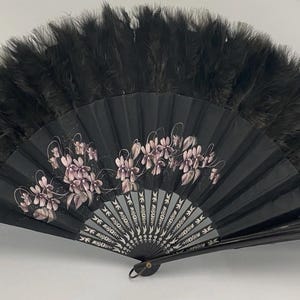Op de afbeelding: Een zwarte handventilator met een bloemenontwerp en zwarte veren. De ventilator heeft een zich herhalend bloemenpatroon in roze en wit. De ventilator is open en toont zijn volledige vorm.