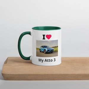 May include: White ceramic mug with a green interior and handle. The mug features the text "I ♥ My Atto 3" with a picture of a blue car. The mug is sitting on a wooden surface.