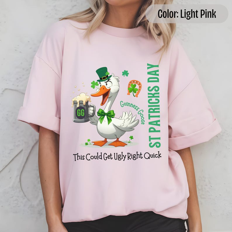A light pink t-shirt featuring a cartoon goose dressed for St. Patricks Day with a green bow tie, leprechaun hat, and shamrocks. The goose, named Guinness Goose, holds a frothy beer mug labeled GG with a playful, mischievous expression. Surrounding elements include a horseshoe and festive decorations. The words ST PATRICKS DAY are written vertically on the right, with the words This Could Get Ugly Right Quick is written horizontally below.