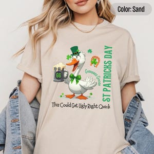 A sand colored t-shirt featuring a cartoon goose dressed for St. Patricks Day with a green bow tie, leprechaun hat, and shamrocks. The goose, named Guinness Goose, holds a frothy beer mug labeled GG with a playful, mischievous expression. Surrounding elements include a horseshoe and festive decorations. The words ST PATRICKS DAY are written vertically on the right, with the words This Could Get Ugly Right Quick is written horizontally below.