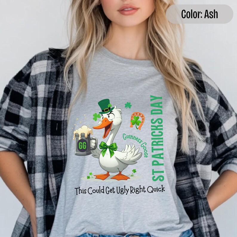 A light grey t-shirt featuring a cartoon goose dressed for St. Patricks Day with a green bow tie, leprechaun hat, and shamrocks. The goose, named Guinness Goose, holds a frothy beer mug labeled GG with a playful, mischievous expression. Surrounding elements include a horseshoe and festive decorations. The words ST PATRICKS DAY are written vertically on the right, with the words This Could Get Ugly Right Quick is written horizontally below.