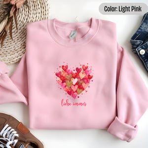 May include: Light pink sweatshirt with a watercolor heart design made of many small hearts in shades of red, pink, and gold. The text "liebe immer" is printed below the heart.