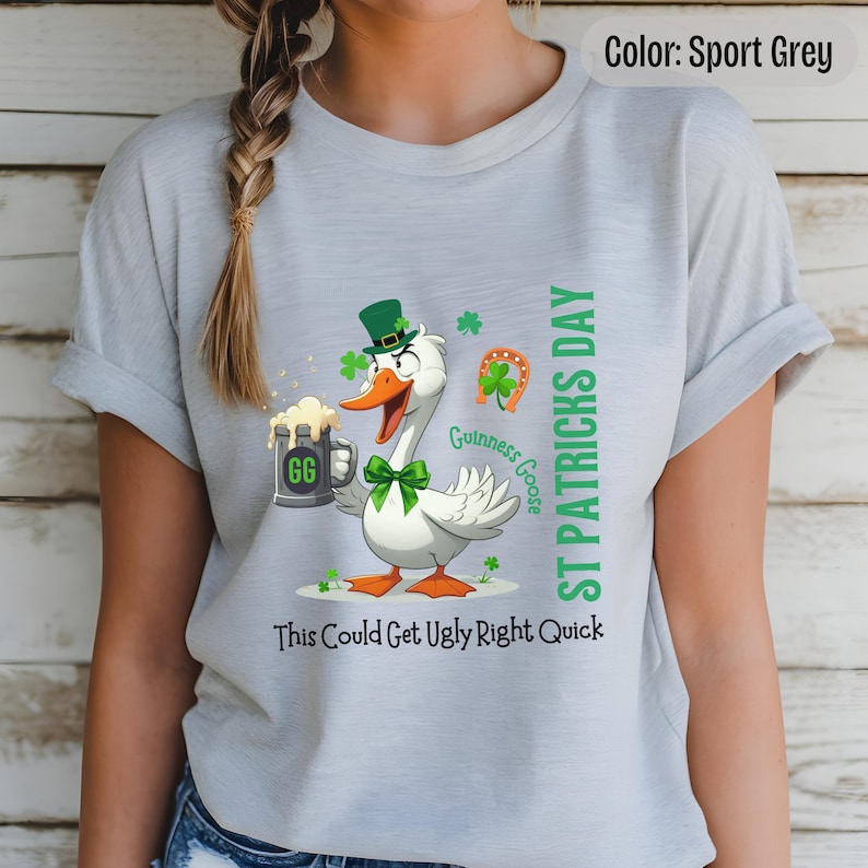 A sport grey t-shirt featuring a cartoon goose dressed for St. Patricks Day with a green bow tie, leprechaun hat, and shamrocks. The goose, named Guinness Goose, holds a frothy beer mug labeled GG with a playful, mischievous expression. Surrounding elements include a horseshoe and festive decorations. The words ST PATRICKS DAY are written vertically on the right, with the words This Could Get Ugly Right Quick is written horizontally below.