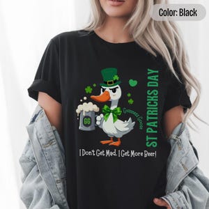 A blak t-shirt featuring a cartoon goose dressed for St. Patricks Day with a green bow tie, leprechaun hat, and shamrocks. The goose, named Guinness Goose, holds a frothy beer mug labeled GG with a playful, mischievous expression. Surrounding elements include shamrocks. The words ST PATRICKS DAY are written vertically on the right, with the words I Dont Get Mad I Get More Beer!, are written horizontally below.