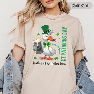 A sand colored t-shirt featuring a cartoon goose dressed for St. Patricks Day with a green bow tie, leprechaun hat, and shamrocks. The goose, named Guinness Goose, holds a frothy beer mug labeled GG with a playful, mischievous expression. Surrounding elements include shamrocks. The words ST PATRICKS DAY are written vertically on the right, with the words How the Heck Am I Getting Home? are written horizontally below.