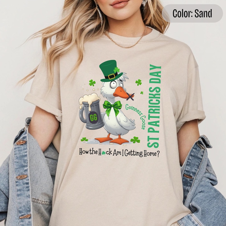 A sand colored t-shirt featuring a cartoon goose dressed for St. Patricks Day with a green bow tie, leprechaun hat, and shamrocks. The goose, named Guinness Goose, holds a frothy beer mug labeled GG with a playful, mischievous expression. Surrounding elements include shamrocks. The words ST PATRICKS DAY are written vertically on the right, with the words How the Heck Am I Getting Home? are written horizontally below.