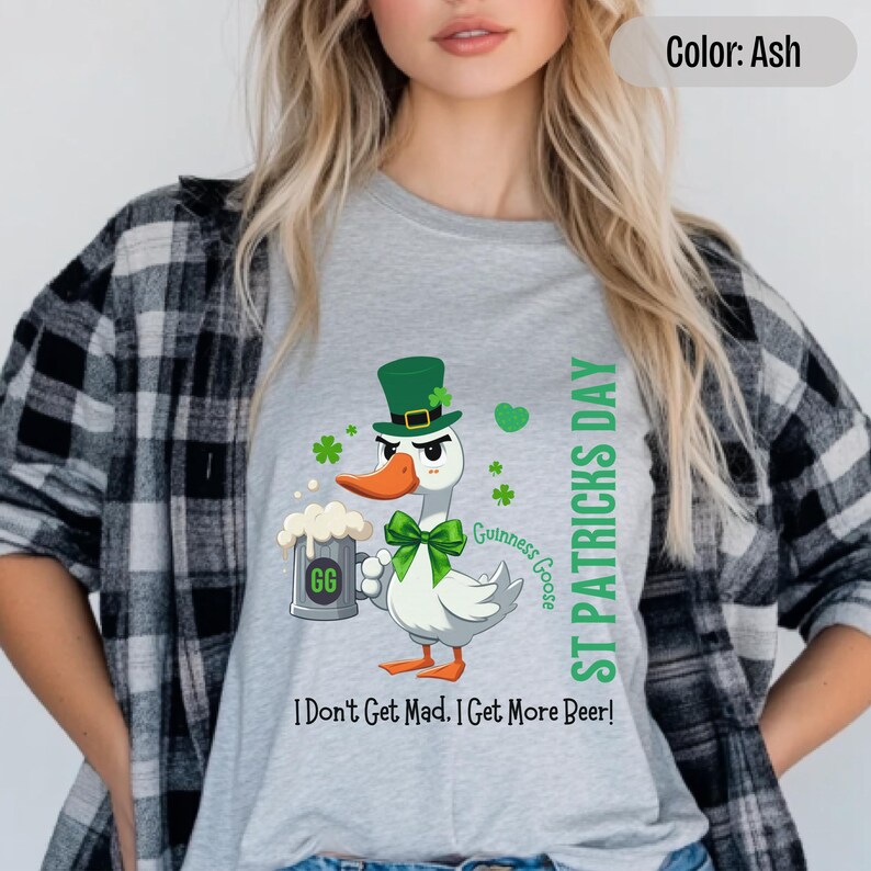 A light grey t-shirt featuring a cartoon goose dressed for St. Patricks Day with a green bow tie, leprechaun hat, and shamrocks. The goose, named Guinness Goose, holds a frothy beer mug labeled GG with a playful, mischievous expression. Surrounding elements include shamrocks. The words ST PATRICKS DAY are written vertically on the right, with the words I Dont Get Mad I Get More Beer!, are written horizontally below.