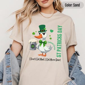 A sand colored t-shirt featuring a cartoon goose dressed for St. Patricks Day with a green bow tie, leprechaun hat, and shamrocks. The goose, named Guinness Goose, holds a frothy beer mug labeled GG with a playful, mischievous expression. Surrounding elements include shamrocks. The words ST PATRICKS DAY are written vertically on the right, with the words I Dont Get Mad I Get More Beer!, are written horizontally below.