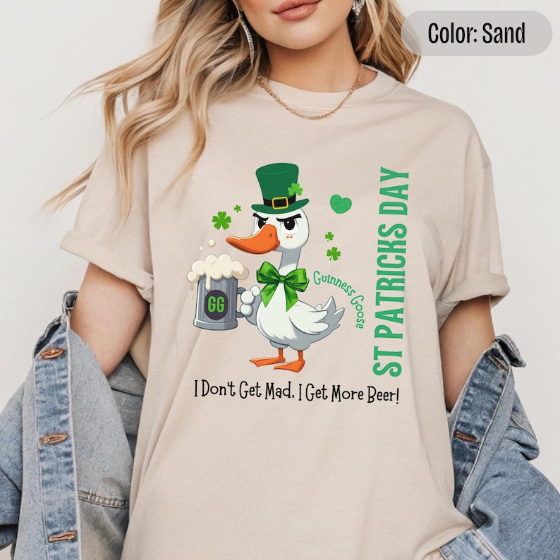 A sand colored t-shirt featuring a cartoon goose dressed for St. Patricks Day with a green bow tie, leprechaun hat, and shamrocks. The goose, named Guinness Goose, holds a frothy beer mug labeled GG with a playful, mischievous expression. Surrounding elements include shamrocks. The words ST PATRICKS DAY are written vertically on the right, with the words I Dont Get Mad I Get More Beer!, are written horizontally below.