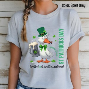 A sport grey t-shirt featuring a cartoon goose dressed for St. Patricks Day with a green bow tie, leprechaun hat, and shamrocks. The goose, named Guinness Goose, holds a frothy beer mug labeled GG with a playful, mischievous expression. Surrounding elements include shamrocks. The words ST PATRICKS DAY are written vertically on the right, with the words How the Heck Am I Getting Home? are written horizontally below.