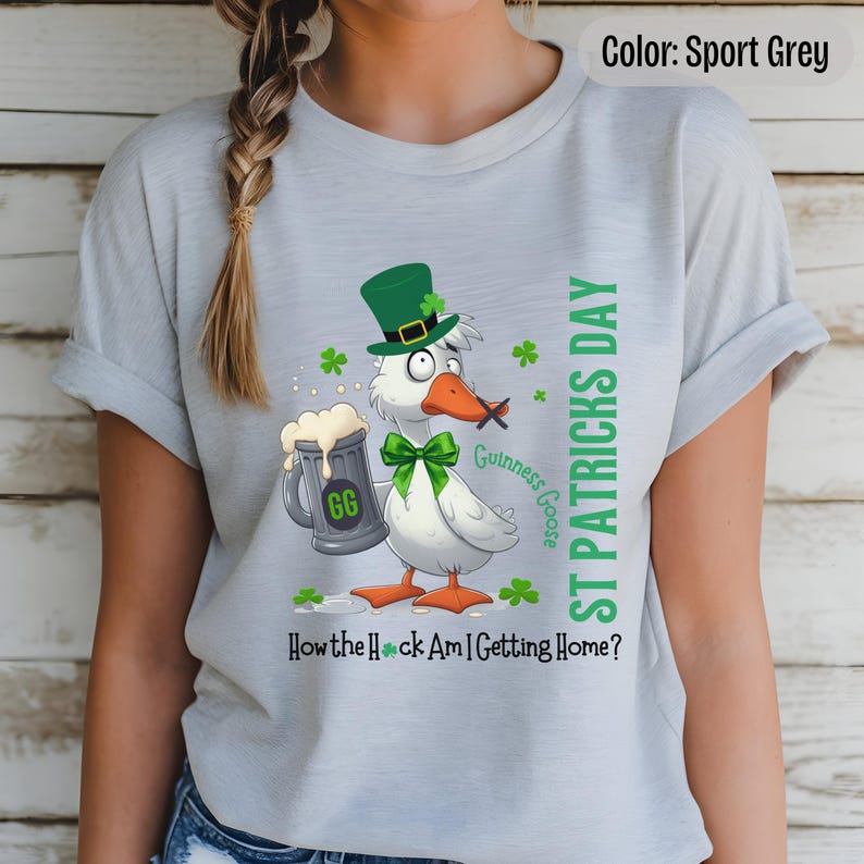 A sport grey t-shirt featuring a cartoon goose dressed for St. Patricks Day with a green bow tie, leprechaun hat, and shamrocks. The goose, named Guinness Goose, holds a frothy beer mug labeled GG with a playful, mischievous expression. Surrounding elements include shamrocks. The words ST PATRICKS DAY are written vertically on the right, with the words How the Heck Am I Getting Home? are written horizontally below.