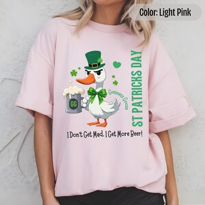 A light pink t-shirt featuring a cartoon goose dressed for St. Patricks Day with a green bow tie, leprechaun hat, and shamrocks. The goose, named Guinness Goose, holds a frothy beer mug labeled GG with a playful, mischievous expression. Surrounding elements include shamrocks. The words ST PATRICKS DAY are written vertically on the right, with the words I Dont Get Mad I Get More Beer!, are written horizontally below.