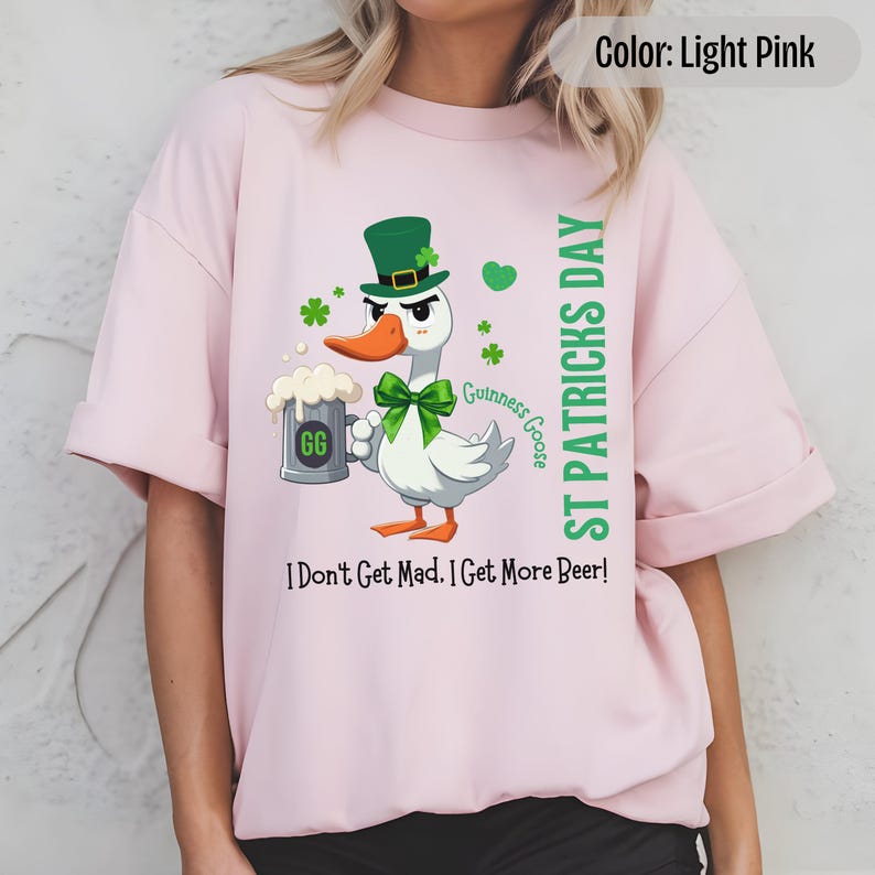 A light pink t-shirt featuring a cartoon goose dressed for St. Patricks Day with a green bow tie, leprechaun hat, and shamrocks. The goose, named Guinness Goose, holds a frothy beer mug labeled GG with a playful, mischievous expression. Surrounding elements include shamrocks. The words ST PATRICKS DAY are written vertically on the right, with the words I Dont Get Mad I Get More Beer!, are written horizontally below.